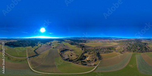 Aerial view of the Seeland District featuring vast agricultural fields and dense forests under a bright sun in Grossaffoltern, Bern, Switzerland.