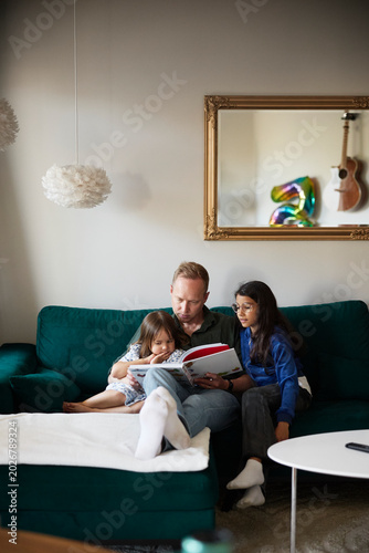 Girls listening to father reading picture book for them while sitting on sofa in living room