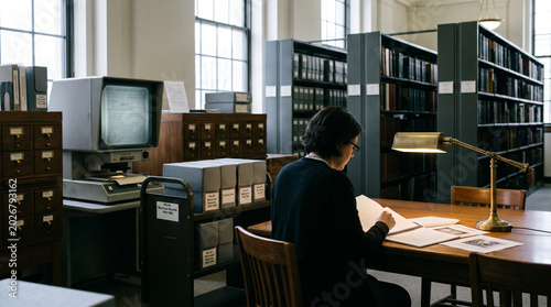A person sits at a table in a library reading a book. Behind them, there are shelves filled with file boxes. An old computer sits on a nearby table. Natural light comes through the windows.