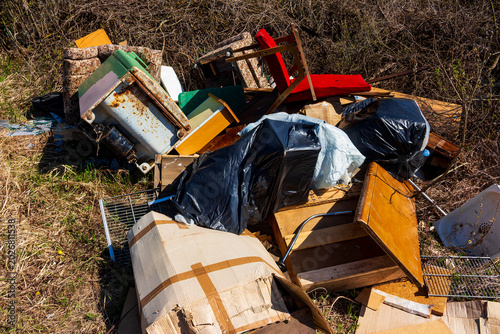 Discarded vintage furniture and trash piled up in a rural field, showing an abandoned heap of weathered household items and cluttered debris amidst overgrown wild bushes and dry grass