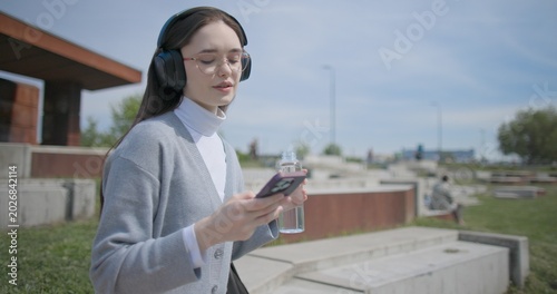 Fotografía A hand-held shot of a young female student in casual clothes, wearing glasses and headphones, sitting on a concrete bench in an urban park, listening to music and drinking water from a bottle