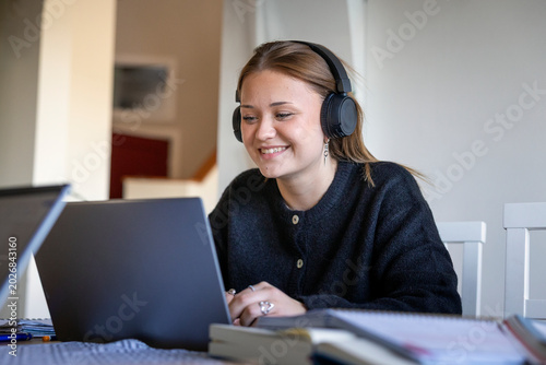 Smiling teenage girl wearing wireless headphones attending online lecture through laptop at home