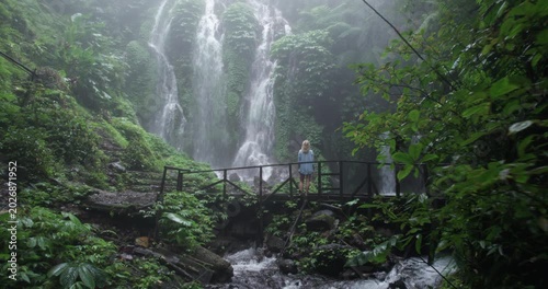 Drone shot of a blonde woman when she stands and admires the view of a waterfall in Bali jungle