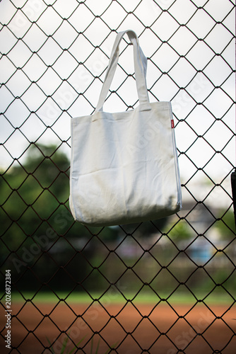 White canvas tote hangs behind chain-link fence. Soft light filters through metal grid pattern. Background shows blurred green foliage and sky. Image evokes minimalist, urban, or eco-conscious vibe