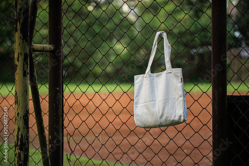 White canvas tote hangs behind chain-link fence. Soft light filters through metal grid pattern. Background shows blurred green foliage and sky. Image evokes minimalist, urban, or eco-conscious vibe