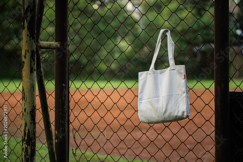White canvas tote hangs behind chain-link fence. Soft light filters through metal grid pattern. Background shows blurred green foliage and sky. Image evokes minimalist, urban, or eco-conscious vibe
