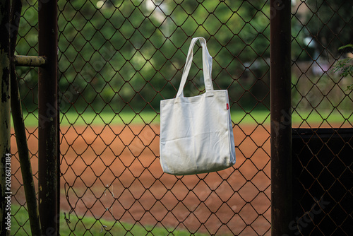 White tote bag hangs on chain-link fence. Soft lighting highlights fabric texture and simple design. Background shows grassy field and blurred trees. Casual