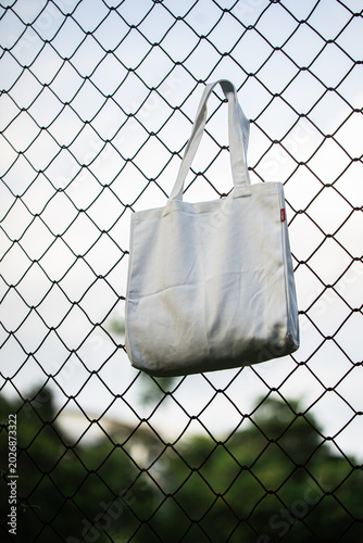 White tote bag hangs on chain-link fence. Soft lighting highlights fabric texture and simple design. Background shows grassy field and blurred trees. Casual