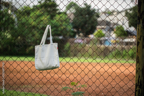 White tote bag hangs on chain-link fence. Soft lighting highlights fabric texture and simple design. Background shows grassy field and blurred trees. Casual