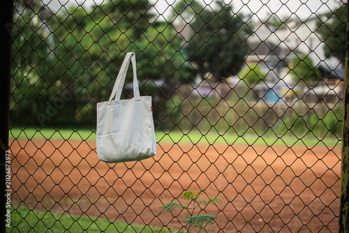 White tote bag hangs on chain-link fence. Soft sunlight highlights fabric's gentle folds. Background features blurred green grass and distant trees. No people visible, suggesting quiet, empty scene