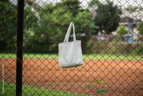 White tote bag hangs on chain-link fence. Soft sunlight highlights fabric's gentle folds. Background features blurred green grass and distant trees. No people visible, suggesting quiet, empty scene