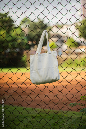 White tote bag hangs on chain-link fence. Soft sunlight highlights fabric's gentle folds. Background features blurred green grass and distant trees. No people visible, suggesting quiet, empty scene