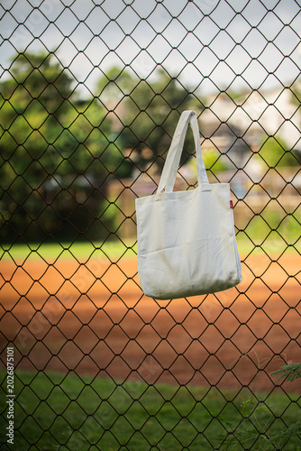 Simple cotton tote hangs on chain-link fence. Sunlight glows behind green trees and grass. Tote shows soft folds and clean lines. Natural scene suggests casual, everyday use