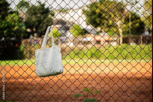 Simple cotton tote hangs on chain-link fence. Sunlight glows behind green trees and grass. Tote shows soft folds and clean lines. Natural scene suggests casual, everyday use
