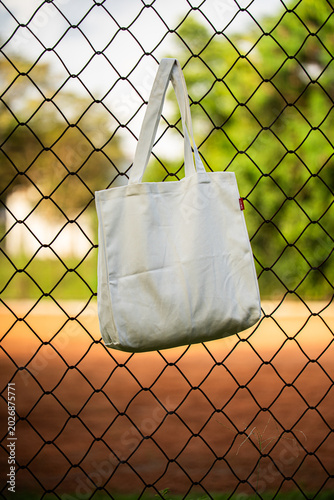 Simple cotton tote hangs on chain-link fence. Sunlight glows behind green trees and grass. Tote shows soft folds and clean lines. Natural scene suggests casual, everyday use