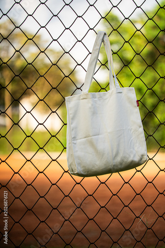 White tote bag hangs on chain-link fence. Blurry background shows green grass and dirt field. Child in distance wears cap and holds bat. Sunlit scene suggests casual park or baseball setting