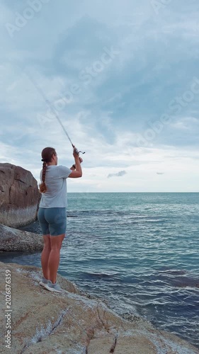 Vertical video. Young Woman Casting Fishing Rod on Rocky Coastline at Dusk, side view of female angler holding rod above sea with wide angle perspective, peaceful Outdoor Recreation