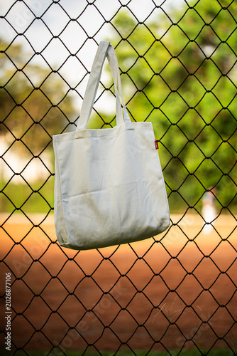 White tote bag hangs on chain-link fence. Blurry background shows green grass and dirt field. Child in distance wears cap and holds bat. Sunlit scene suggests casual park or baseball setting