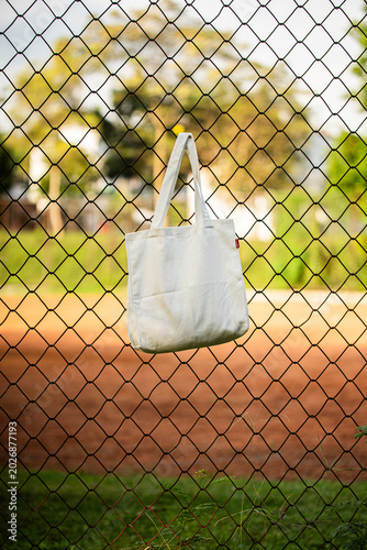 Simple tote bag hangs on chain-link fence. Soft fabric shows subtle wear and natural folds. Background holds green trees and blurred backyard. Scene suggests casual, everyday outdoor setting