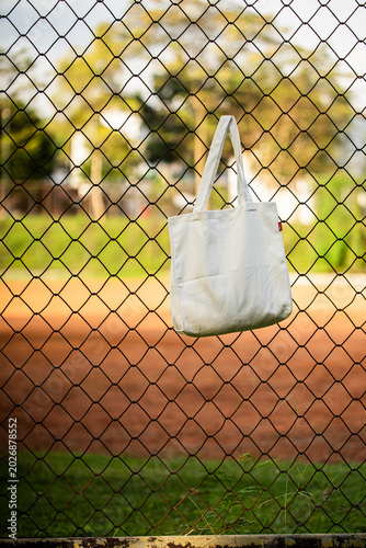 Simple tote bag hangs on chain-link fence. Soft fabric shows subtle wear and natural folds. Background holds green trees and blurred backyard. Scene suggests casual, everyday outdoor setting