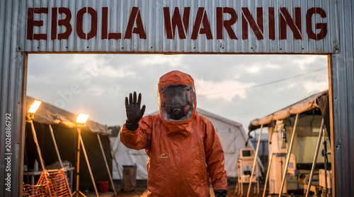 Worker in orange hazmat suit standing under ebola warning sign outdoors