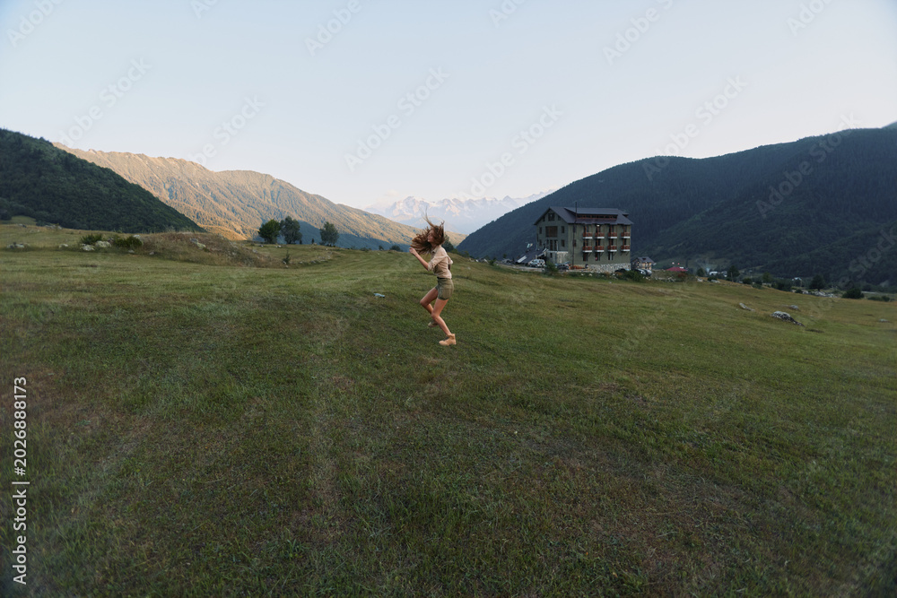 Fototapeta premium Young woman jumps joyfully on green grass field in mountain valley near house during sunny day, enjoying active lifestyle and nature outdoors.