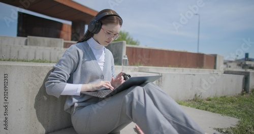Fotografija A hand-held shot of a young female student in casual clothes, glasses, and headphones working and typing on a laptop while sitting on a concrete bench in an urban park