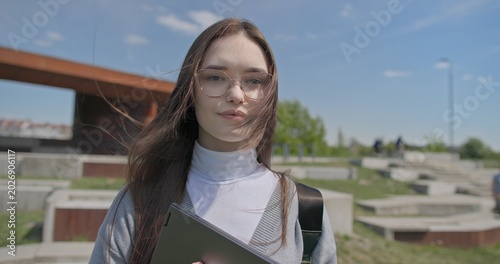 A hand-held portrait of a young female student in casual clothes, wearing glasses and a backpack, holding a notebook and laptop in her hands and posing looking at the camera while standing in a park 
