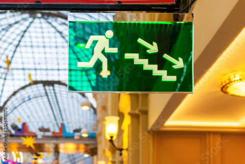 Green emergency exit sign indicating stairs down in a public shopping mall. Fire safety information board and evacuation route illumination for building security.