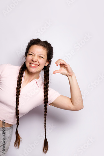 Happy young woman with long braids posing in studio. Cheerful girl making heart shape hand gesture, winking and smiling. Beautiful model in pink polo t-shirt isolated on white background