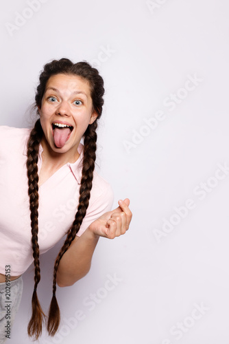 Funny young woman with long braids showing tongue. Cheerful girl making money hand gesture, winking and grimacing. Emotional model in pink polo shirt posing studio portrait on white background