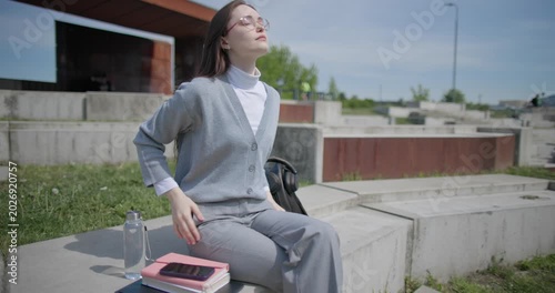 A handheld shot of a young female student in casual clothes sitting on a concrete bench in an urban park, enjoying a sunny summer day.