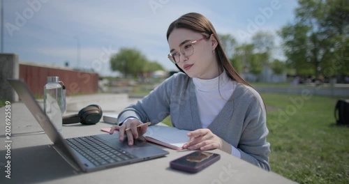 A hand-held shot of a young female student in casual clothes and glasses using a laptop and taking notes with a pen in a notebook in an urban park.