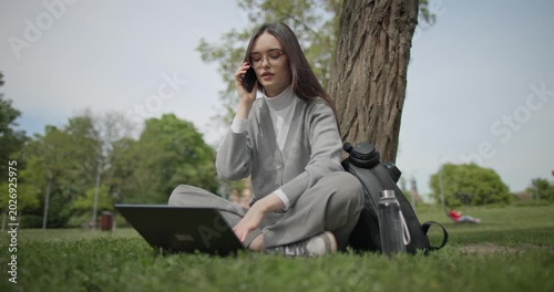 A hand-held shot of a young, beautiful female student in casual clothes, wearing glasses and headphones, sitting on the grass under a tree in a city park, using a laptop and talking on a mobile phone.
