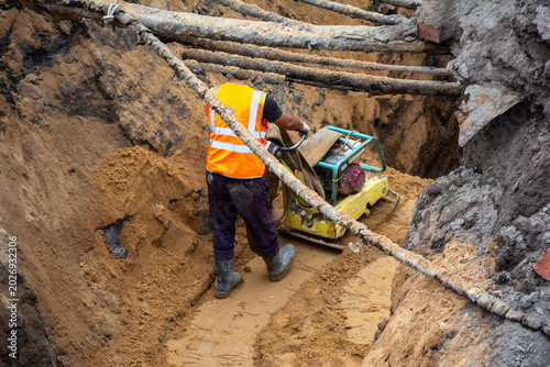 Construction worker in safety vest operating a compactor machine in a trench filled with sand, surrounded by wooden supports and earth walls