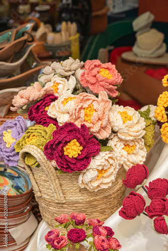 Crochet flower bouquet fills woven basket at craft stall, with handmade yarn roses and blossoms among ceramics. Concept of crochet decor, handmade gift, artisan market, hobby craft, textile design