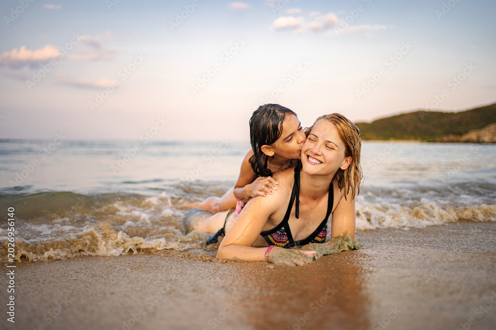 Fototapeta premium Mother and daughter smiling, lying in waves on a sandy beach
