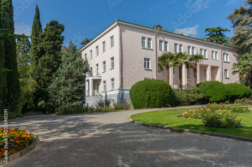 Bild auf Leinwand Two-story light pink building with white pillars and numerous windows, surrounded by greenery including palm trees, trimmed bushes and flowering plants