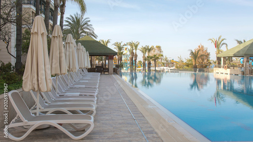 Luxury tropical resort with large infinity swimming pool, white sun loungers and umbrellas lined beside crystal clear water among palm trees, peaceful summer vacation destination concept