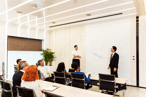 Professionals attend a business presentation in a modern boardroom while two presenters explain charts and strategy on a whiteboard