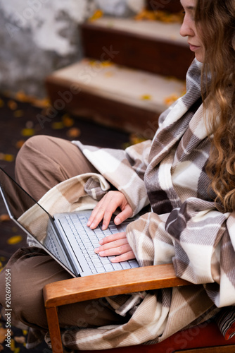 A young curly-haired woman is working on a laptop outside her home during the autumn season. She is wearing a casual outfit and plaid.
