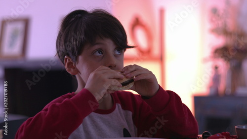 Photos Young boy eating hamburger at dining table, taking big bite during indoor family