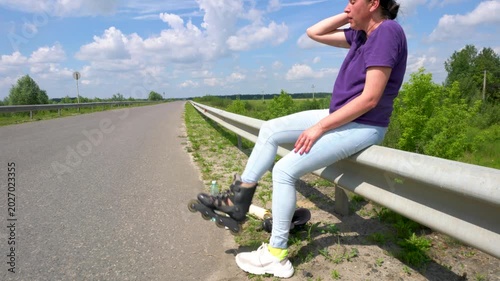 An adult woman in jeans takes off her roller skates on an asphalt road.