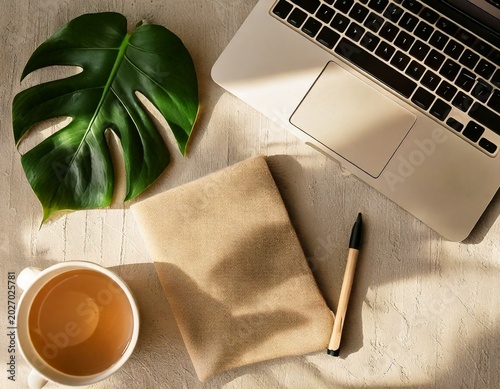 Modern flat lay of a beige laptop, coffee mug, notebook, pen, and green leaf on a textured surface, perfect for a serene workspace.