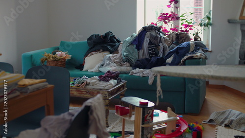 Massive stack of unfolded clothes covering living room sofa and spilling onto cushions highlighting domestic overload and never ending household responsibilities