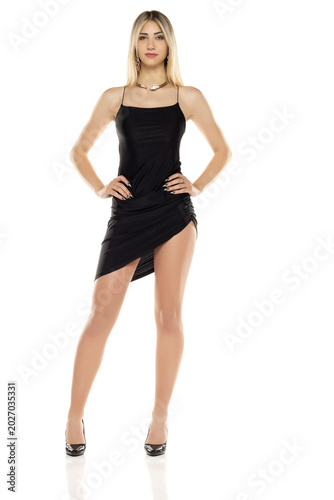 Full length studio shot of blonde woman in short black asymmetrical dress and high heels posing confidently on white background