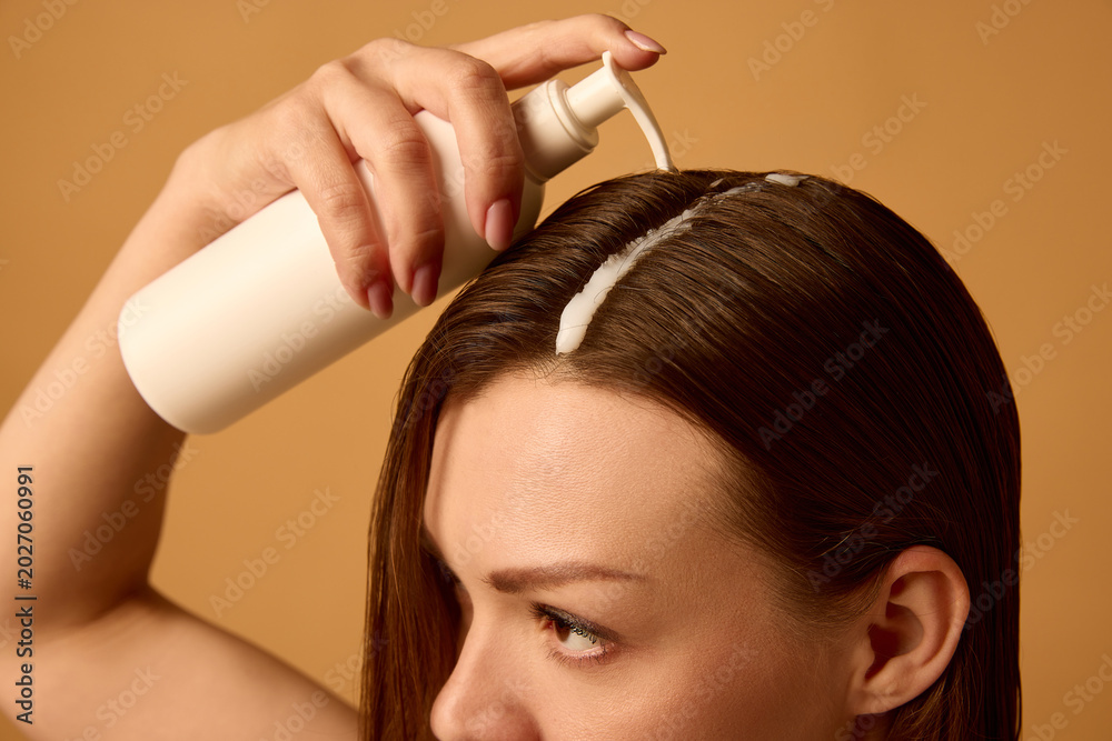 Fototapeta premium Woman applying hair serum from pump bottle to wet brunette hair on warm studio background. Concept of scalp treatment, leave-in care, clean beauty promo, hydration and repair.