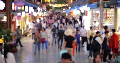 Blurred view of citizens strolling along wide pedestrian shopping street in downtown Kunming, passing brightly lit retail storefronts and marketplace stalls with neat civil twilight adding to scene.
