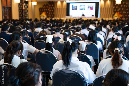 Large group of diverse students in uniforms sitting in a grand convention hall listening to an educational seminar with a speaker on stage, authentic academic conference and learning concept