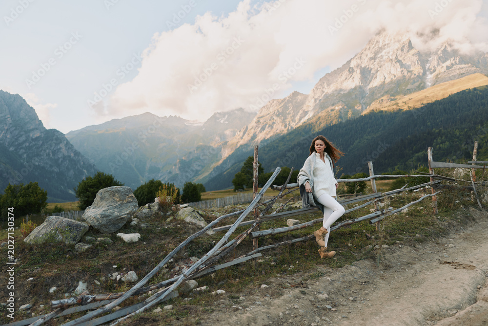 Fototapeta premium Young woman in white outfit leans on rustic wooden fence with mountain landscape in the background during a clear day.
